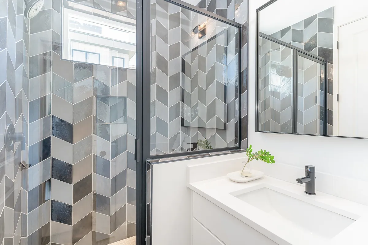 A bathroom showcasing a stylish hexagonal tile shower wall with black-framed glass, a white sink with a modern faucet, and a well-lit mirror. The design is fresh and rejuvenating, blending modern fixtures with comforting touches.