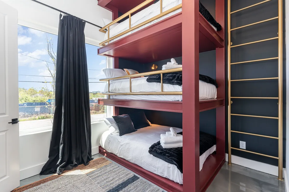 A cheerful kid-friendly bunk room with two bunk beds, a large window, black-out curtains, and a soft rug. The space is inviting and playful, ideal for young guests to sleep, play, or read.