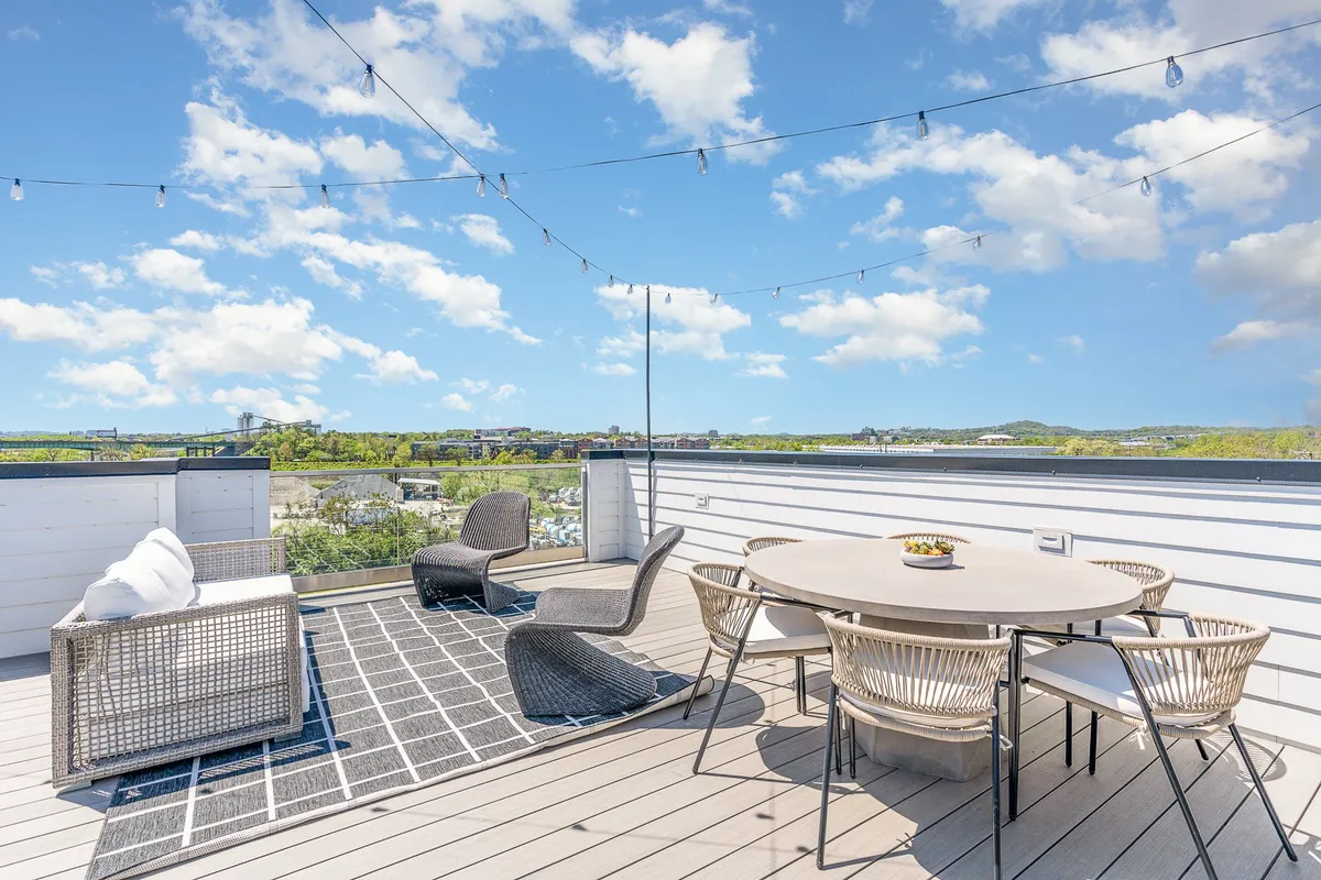 A spacious rooftop deck is shown at sunset, furnished with a large outdoor dining table, lounge chairs, and illuminated by string lights. The vantage point offers a scenic view of the surrounding city and skyline, creating an ideal setting for al fresco meals and evening gatherings.