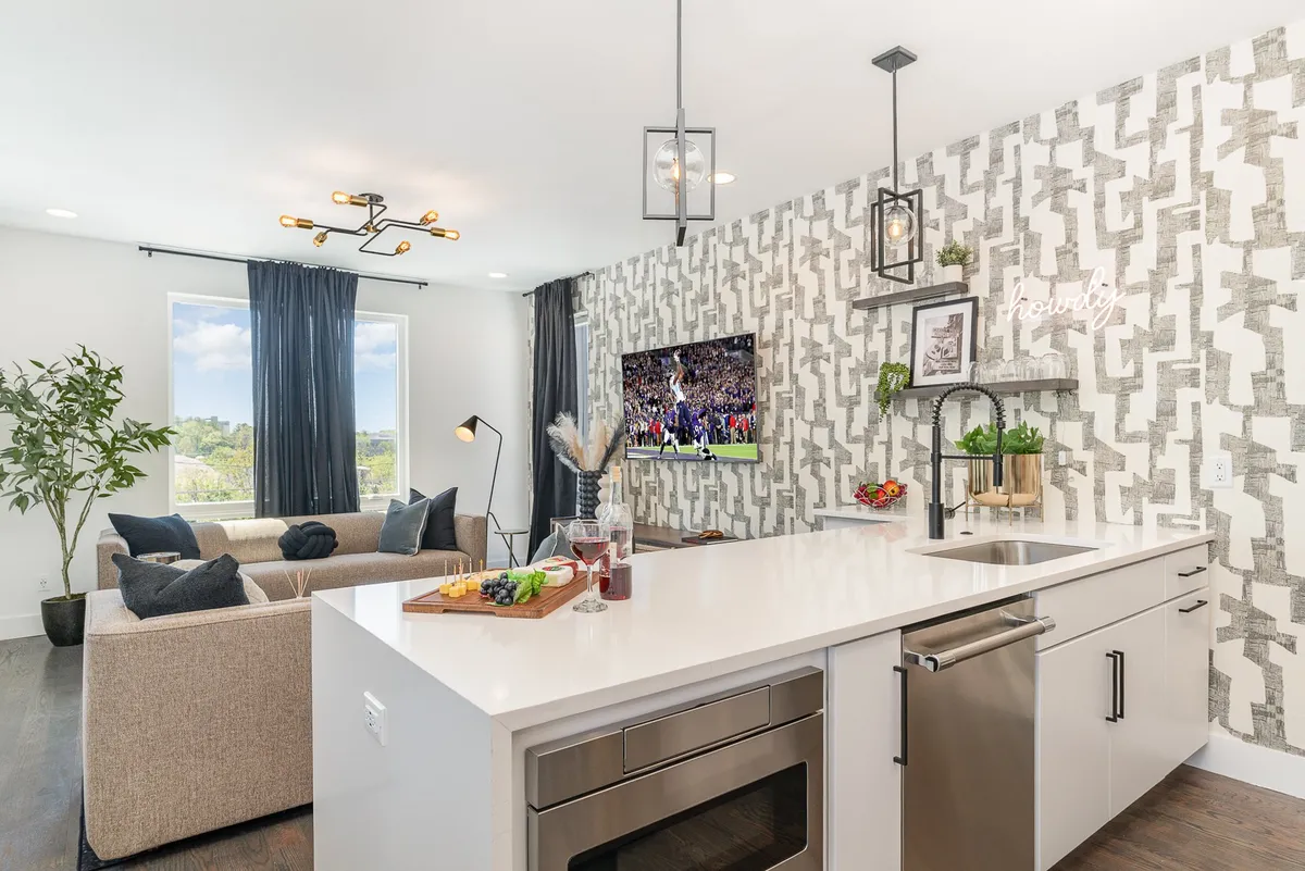 A welcoming shot of the kitchen island open to a sofa and TV area. The image highlights the flow between cooking, dining, and relaxing spaces, with premium appliances and stylish décor touches clearly visible. Plants and a decorative wall add warmth and vibrancy.