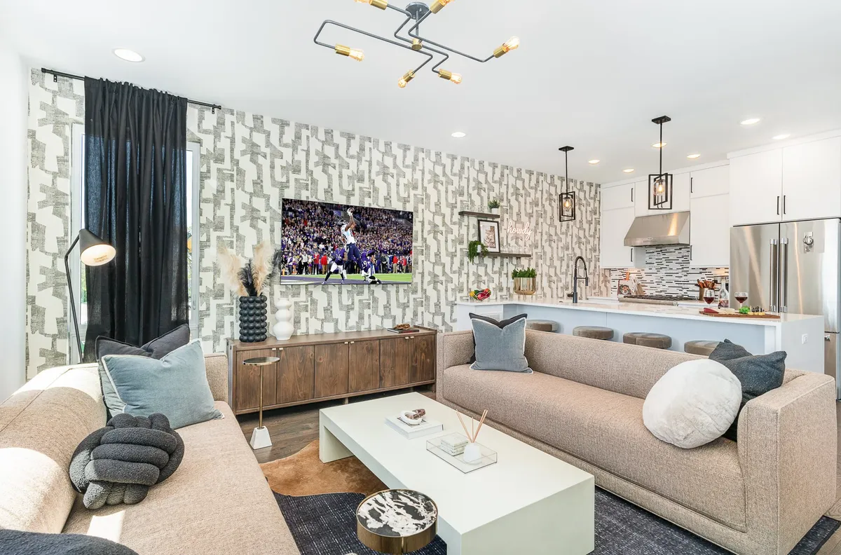 This image captures an open-concept interior featuring a spacious seating area with sofas and a coffee table, a large television displaying a sports event, a decorative wall, and a visible kitchen area in the background. Generous windows let in natural light, and the layout encourages social interaction and relaxation.