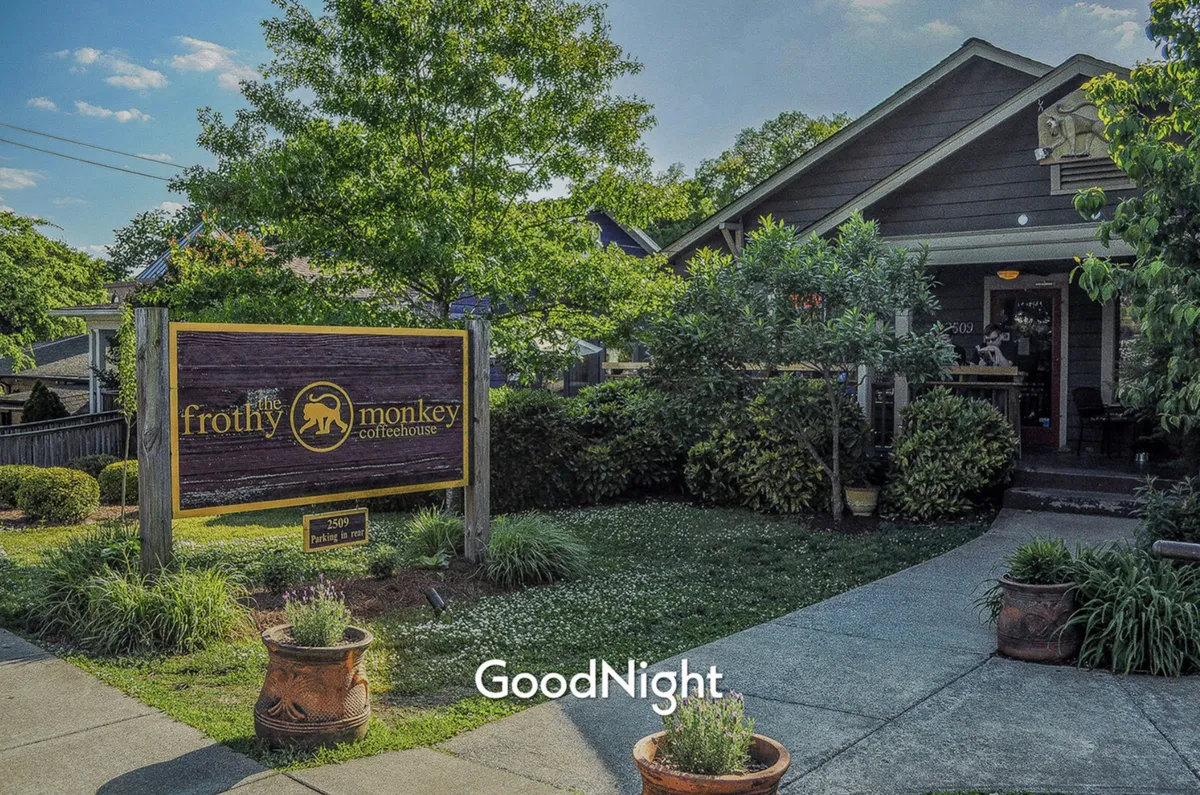 This photo focuses on the Frothy Monkey coffeehouse entrance, adorned with its iconic sign, surrounded by greenery, and featuring an inviting patio area. The ambiance is warm and relaxed, with visible cues of people enjoying drinks and casual conversation.