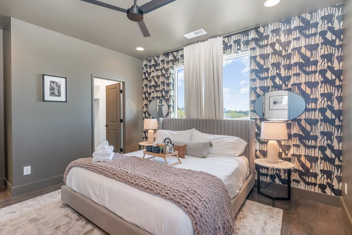 A warmly lit bedroom featuring a plush bed with soft bedding, decorative pillows, nightstands with lamps, artwork on the wall, and large windows draped with curtains. The room exudes a sense of coziness and tranquility, ideal for rest and relaxation.