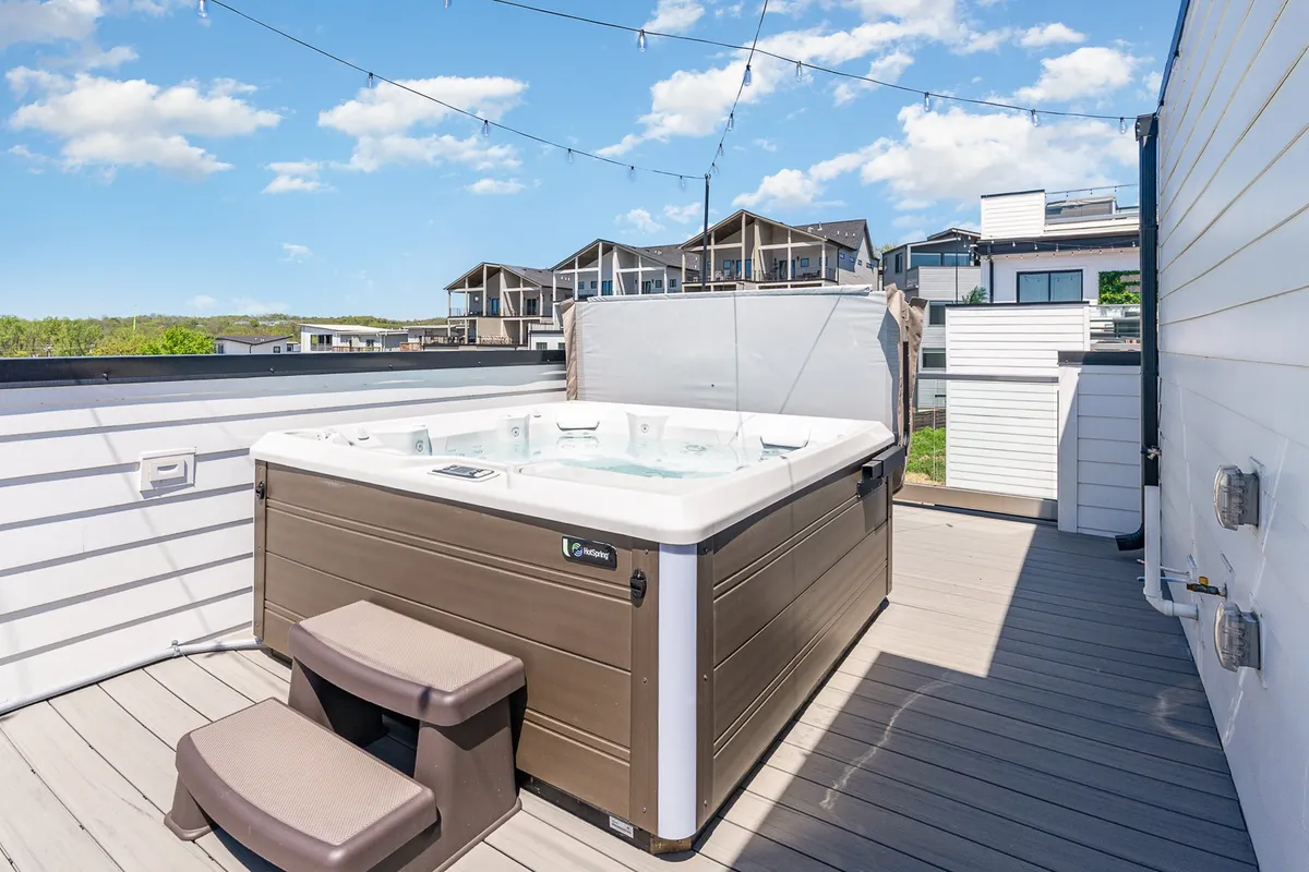 This image captures a stylish rooftop deck featuring a bubbling hot tub, cozy seating areas, and warm bistro string lights overhead. The city skyline is visible in the background, and the space exudes an inviting, intimate atmosphere perfect for evening relaxation or social gatherings.