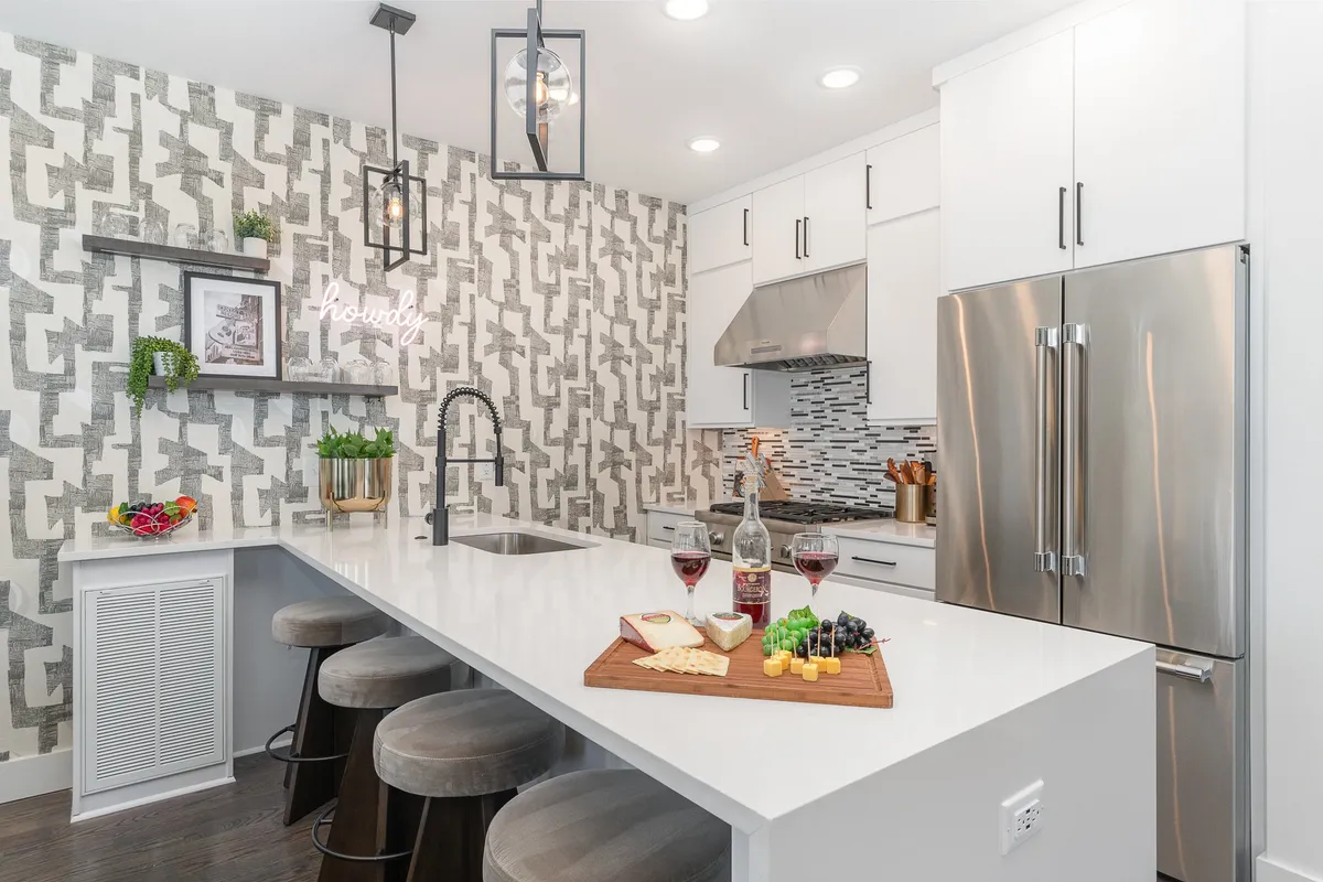 A stylish kitchen scene with an island countertop set for entertaining—wine and cheese arranged artfully, modern light fixtures overhead, and textured wallpaper adding flair. The inviting setup suggests both culinary creativity and social gatherings.